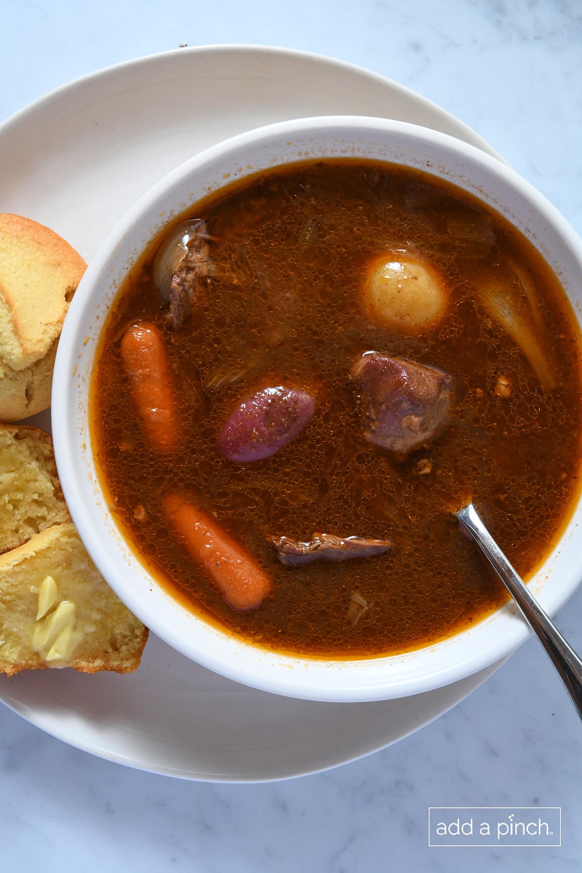 Soup bowl filled with venison stew on a plate with buttered cornbread muffin.